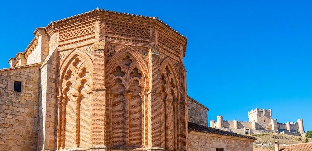 Plano medio de la Iglesia de San Pablo en Peñafiel, resaltando su espectacular ábside de estilo gótico-mudéjar construido en ladrillo rojizo con arcos lobulados. Al fondo, dominando la composición sobre el cerro, se alza imponente la silueta del Castillo de Peñafiel (Museo del Vino) bajo un cielo azul intenso.