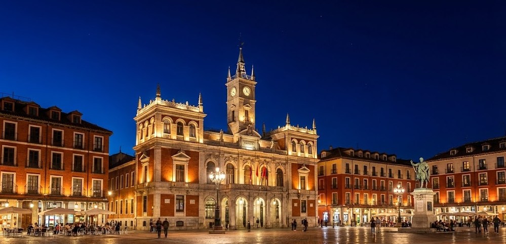 Vista nocturna de la Plaza Mayor de Valladolid, con la iluminación monumental resaltando las fachadas de los edificios y el Ayuntamiento bajo un cielo azul oscuro.