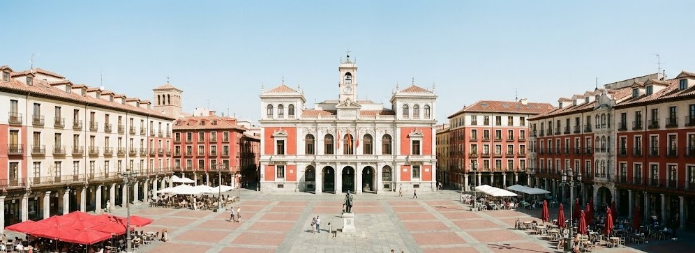 Vista panorámica de la Plaza Mayor de Valladolid mostrando el edificio del Ayuntamiento al fondo con su fachada de color rojo almagre y los portales circundantes.