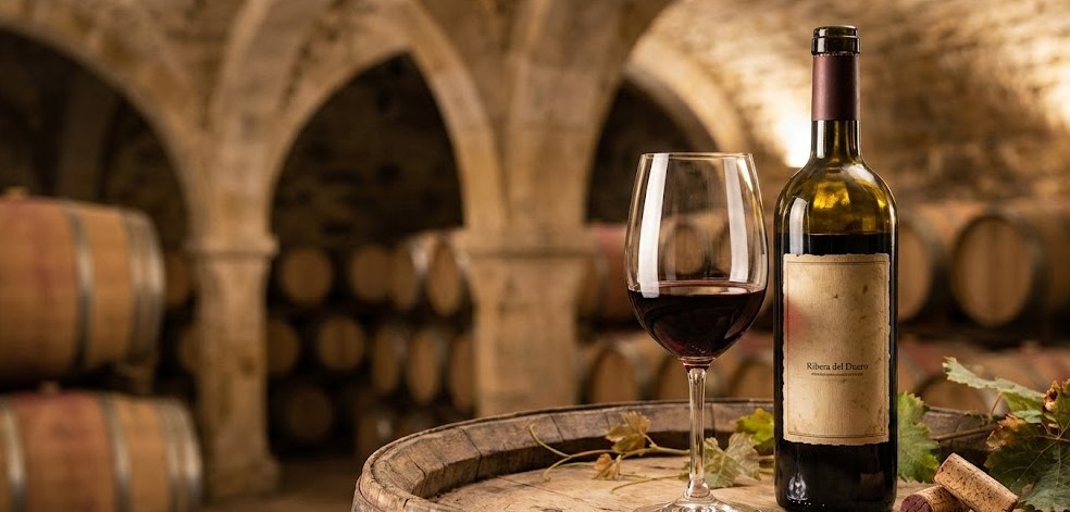 Bodegón elegante y evocador de enoturismo. Primer plano de una copa de cristal con vino tinto Ribera del Duero recién servido, junto a una botella de etiqueta clásica sobre la textura de madera de una barrica de roble. El fondo muestra desenfocados los arcos de piedra de una antigua bodega subterránea, sugiriendo tradición y calidez.